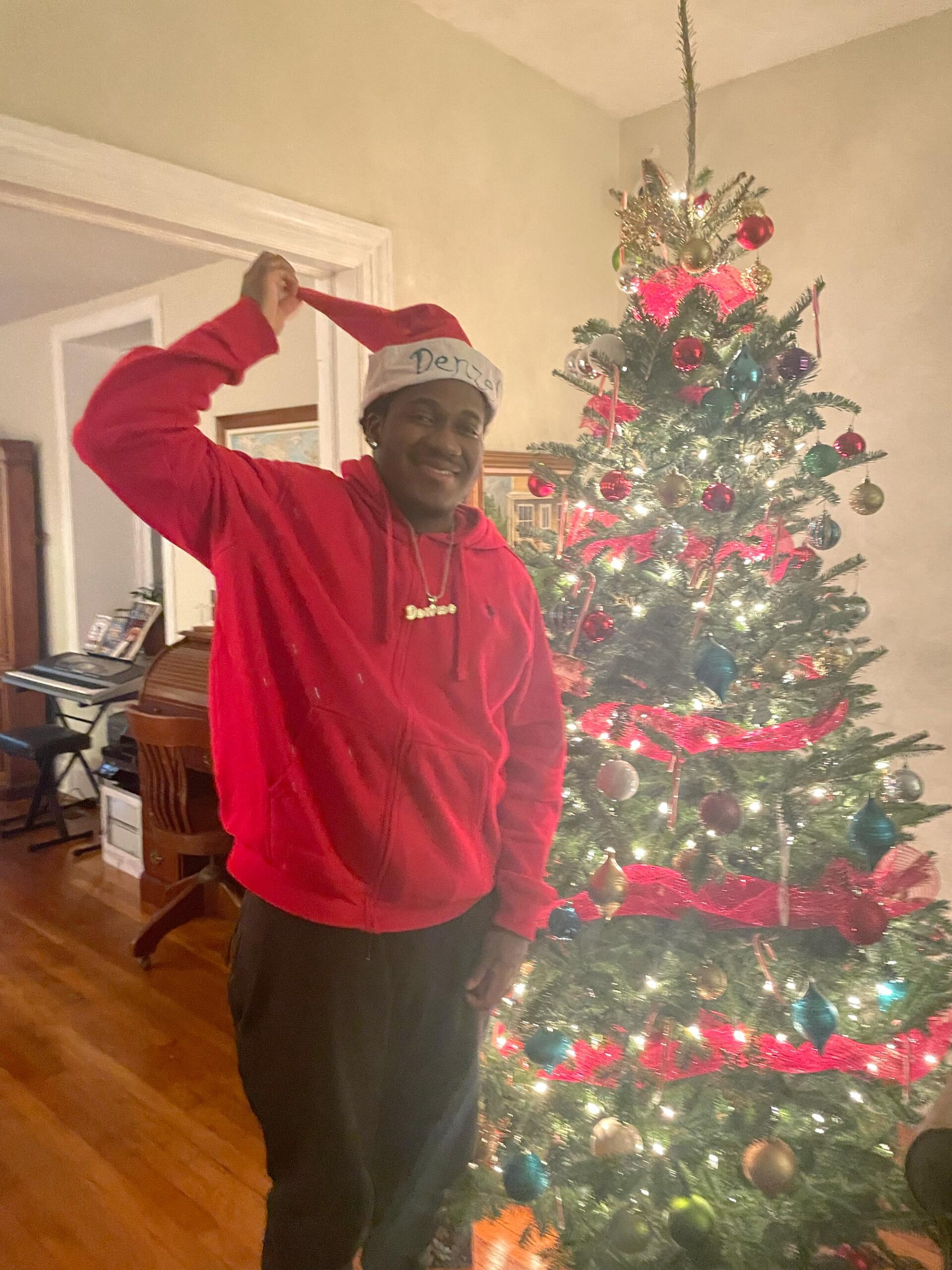 Smiling black man wearing a red hoodie, black pants, and a Santa hat that reads "Denzel", standing in front of a Christmas tree.