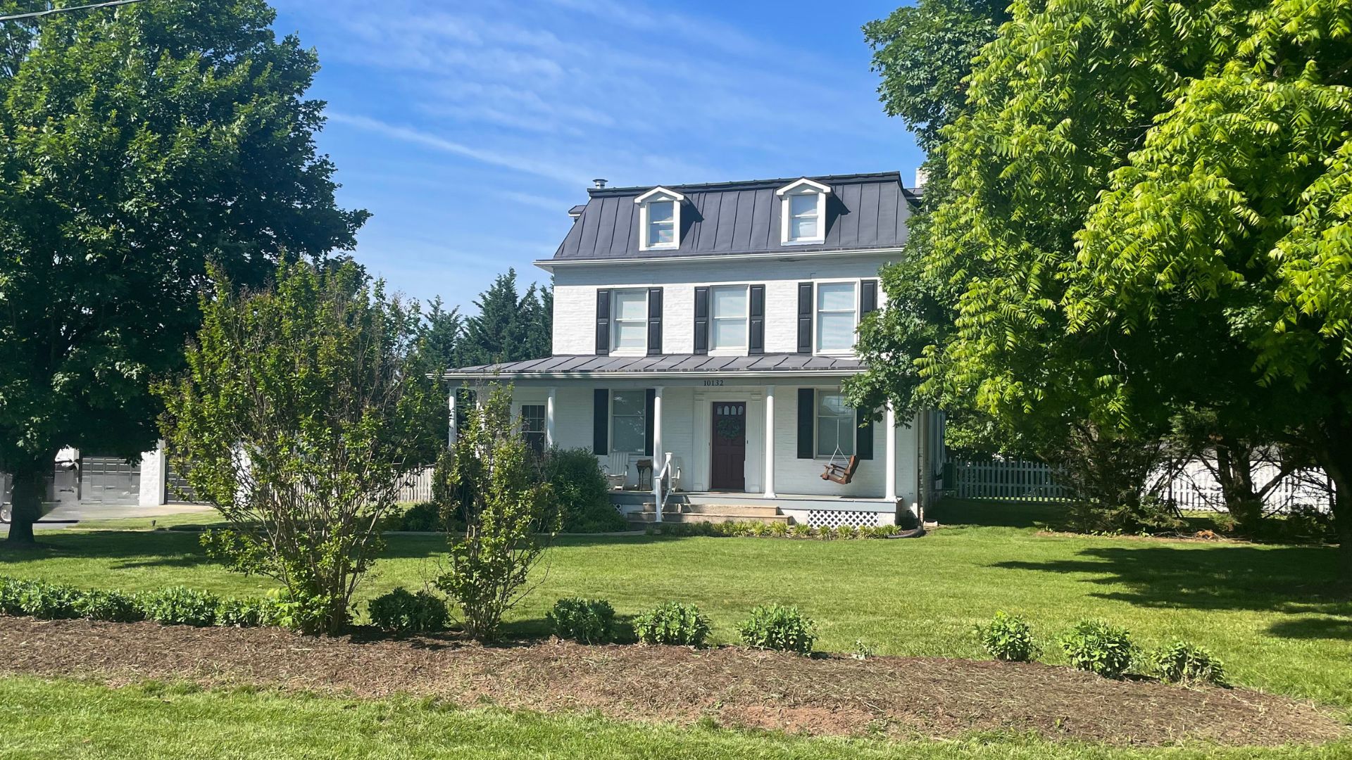 color photo of Steadfast farmhouse in summer with two large full, green trees, green lawn, and surrounding landscaping