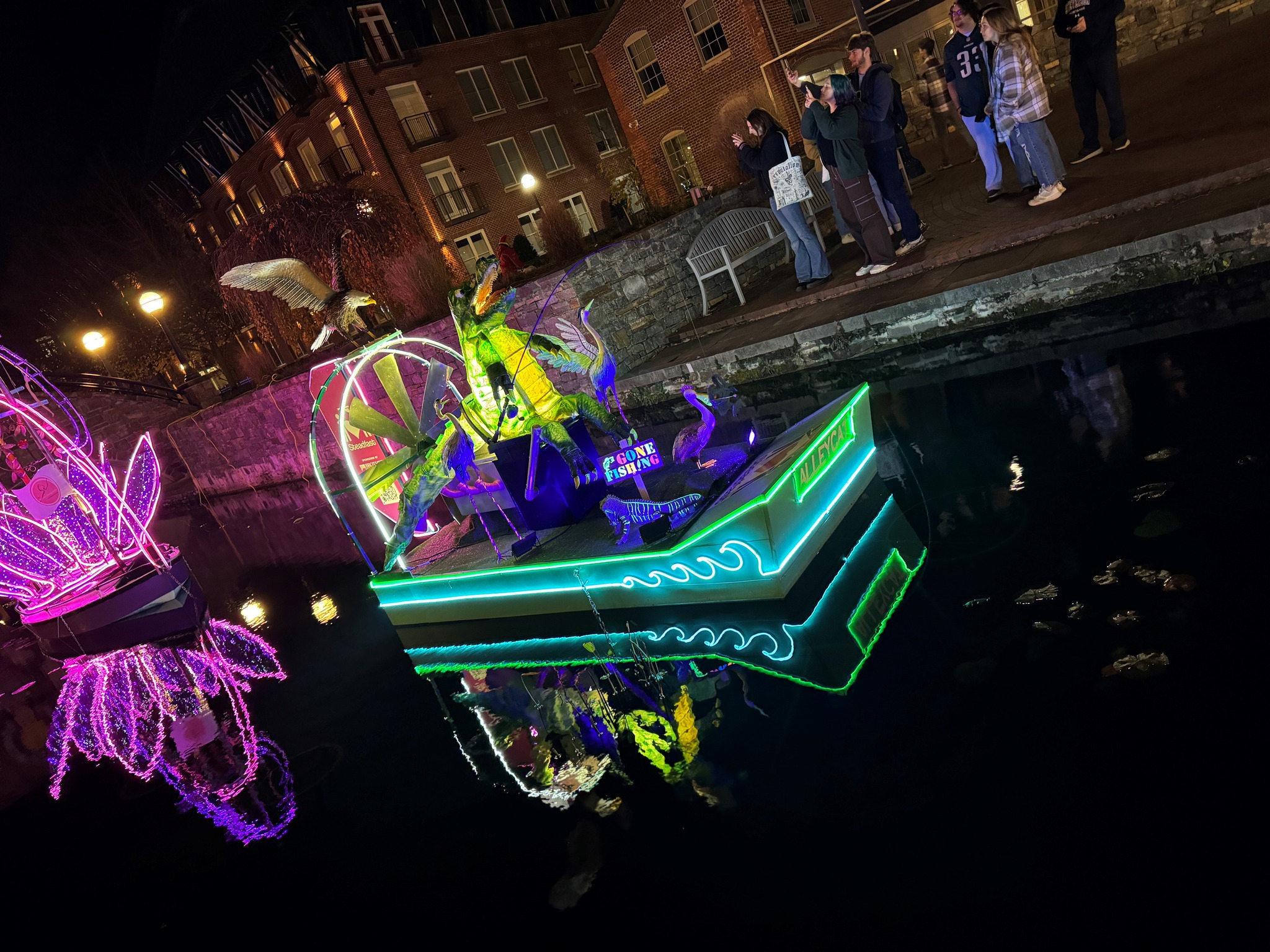 nighttime photo of Steadfast's boat with lights on Carroll Creek for the 2022-23 Sailing Through the Winter Solstice with people on taking pictures from the cobblestone walk lining the creek